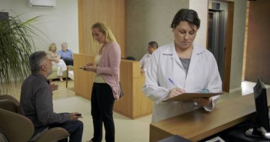 Hastalar arka planda beklerken ve çalışanlar yoğun tıbbi lobide ziyaretçilerle uğraşırken klinik resepsiyonunda not alan kadın doktor.