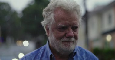 Yağmurda acı tatlı gülümseyen yaşlı bir adam, gözyaşı dolu gözler ve hüznü ve kabulü yansıtan şefkatli bir ifade.