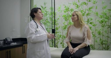Doktor, yeşil bambu bitkisi geçmişi olan klinikteki sağlık danışmanlığı sırasında muayene masasında oturan orta yaşlı kadın hastayla konuşuyor.