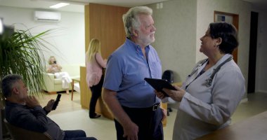Kadın doktoru dinlerken gülümseyen kıdemli adam klinik lobisindeki dijital tablet hakkında bilgi veriyor, resepsiyon masasının yanındaki günlük sağlık danışmanlığı