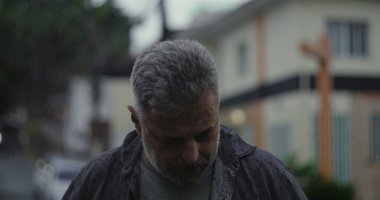 Orta yaşlı bir adam yağmurun altında duruyor. Islak giysiler ve kasvetli bir ifade, bulanık arabalar ve karamsar sokak atmosferiyle çevrili.