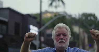 Yaşlı bir adam yağmurda kolları hafifçe havada duruyor, kameraya geniş gözlerle bakıyor ve şok, inançsızlık ve duygusal hassasiyet ifadesi sergiliyor.