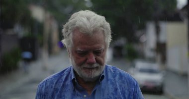 Yaşlı bir adam yağmurun altında üzgün gözlerle ve duygusal ifadeyle, dudakları sımsıkı ve kasvetli bir şekilde duruyor.