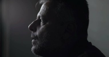 Düşük ışık kliniğindeki orta yaşlı adamın yakın çekimi, düşünceli sükûnetten duygusal sıkıntıya geçiş, teşhis ve içsel turmoilere karşı sessiz tepkiler.