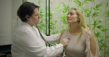 Klinikteki orta yaşlı kadın hastanın kalbini incelemek için steteskop kullanan kadın doktor rutin kontrolleri ve kişiselleştirilmiş tıbbi arabaları vurguluyor.