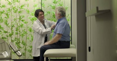 Gülümseyen doktor, klinikteki yaşlı erkek hastaya steteskopla arkadaşça tıbbi muayene yapıyor.