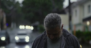 Orta yaşlı bir adam yağmurun altında duruyor. Islak giysiler ve kasvetli bir ifade, bulanık arabalar ve karamsar sokak atmosferiyle çevrili.