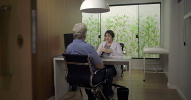 Klinikteki özel danışma sırasında yaşlı erkek hastayla konuşan doktor, profesyonel sağlık hizmetleri interaktifi hakkında yakın perspektif için kapı aralığından çerçevelenmiş