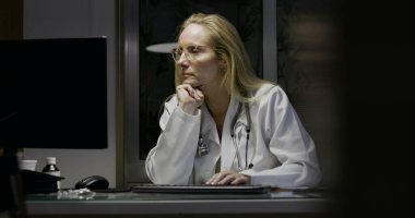 Kadın doktor gece vardiyasında çalışıyor, bilgisayar ekranını izlerken çenesini dinlendiriyor, derin düşüncelere dalmış ve modern klinik setin tıbbi öğeleriyle çevrili.