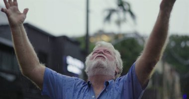 Kolları geniş ve yüzü göğe yükselmiş yaşlı bir adam, ruhsal rahatlama, minnettarlık veya duygusal teslimiyet ifadesiyle yağmuru kucaklıyor.