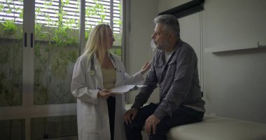 Kadın doktor gülümseyen orta yaşlı erkek hasta ile tıbbi notları tartışırken clini 'de muayene masasında oturuyor.