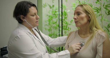 Doktor orta yaşlı kadın hastaların kalp atışlarını dinlemek için steteskop kullanıyor. Klinikte yeşil bitki duvarı arka planına sahip tıbbi muayene sırasında.