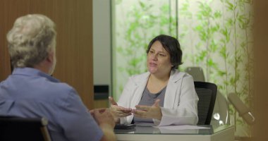 Klinik ofis ortamında kıdemli erkek hastayla yapılan istişare sırasında el hareketleriyle tedavi planını açıklayan doktor