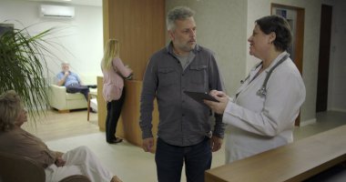 Adam, klinik koridorunda kadın doktorla konuşurken rahatladığını ifade ediyor. Elleri göğsünde gülümsüyor. Sağlık hizmetleri tartışmaları sırasında güven verici haberler alıyor.