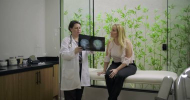 Doktor, klinikte yeşil bambu bitkisi geçmişi olan nörolojik danışma sırasında orta yaşlı bir kadınla beyin röntgenini inceliyor.