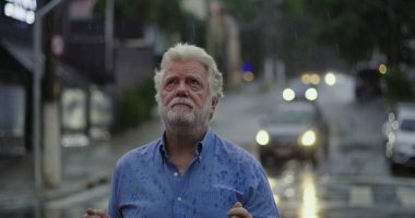 Yağmurda ayakta duran yaşlı adam, elleri açık ve duygusal ifade ile yukarı bakıyor keder, kafa karışıklığı, ya da ruhsal özlem şehir caddesinde