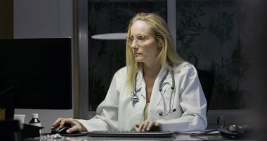 Masada geç saatlere kadar çalışırken not alan kadın doktor, tıbbi malzemeler ve belgelerle çevrili, odaklanmış ifadeli bilgisayar monitöründen uzağa bakıyor.