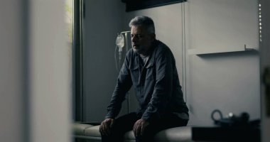 Sıkıntılı adam klinikte yalnız başına oturuyor, ağır duruşuyla öne eğiliyor, bastırılmış ışıklandırma yalnızlığı ve duygusal çöküntüyü vurguluyor.