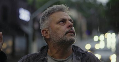 Orta yaşlı bir adam yağmurda dalgın dalgın bir ifadeyle yukarı bakıyor, boş şehir sahasında öylece duruyor.