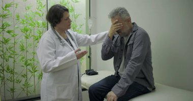 Orta yaşlı erkek hasta sinir bozucu teşhislere tepki verirken, kadın doktor klinik ortamda tıbbi konsültasyon sırasında duygusal destek sunuyor.