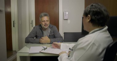 Orta yaşlı hasta klinikteki danışma sırasında doktorunu dikkatle dinliyor. Elinde not ve not defteriyle.