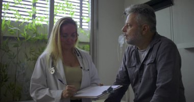 Orta yaşlı erkek hasta tıbbi danışma odasında dikkatle dinlerken kadın doktor panoya yazı yazıyor.