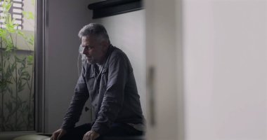 Orta yaşlı bir adam, boş bir muayenehanede gergin bir duruşla öne eğiliyor. Zor bir muayeneden sonra, yüzündeki gerginlik ve yorgunluk görülebiliyor..