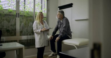 Modern tıp bürosunda muayene masasında oturan erkek hastayla konuşurken not alan kadın doktor, tıbbi danışma süreci ve özenli ifade ile devam ediyor.