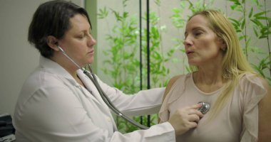 Klinikteki orta yaşlı kadın hastanın kalbini incelemek için steteskop kullanan kadın doktor rutin kontrolleri ve kişiselleştirilmiş tıbbi arabaları vurguluyor.