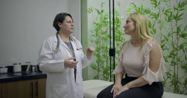 Doktor, yeşil bambu bitkisi geçmişi olan klinikteki sağlık danışmanlığı sırasında muayene masasında oturan orta yaşlı kadın hastayla konuşuyor.