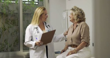 Klinik muayene odasında yaşlı bir kadın hastayla teşhisi tartışırken gülümseyen kadın doktor, her ikisi de arka planda serumla sıcak temas kuruyor.