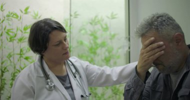Orta yaşlı erkek hasta sinir bozucu teşhislere tepki verirken, kadın doktor klinik ortamda tıbbi konsültasyon sırasında duygusal destek sunuyor.
