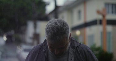 Orta yaşlı bir adam yağmurun altında duruyor. Islak giysiler ve kasvetli bir ifade, bulanık arabalar ve karamsar sokak atmosferiyle çevrili.