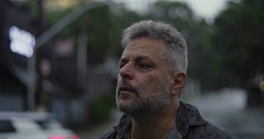 Orta yaşlı bir adam, ciddi bir şekilde aşağıya bakıyor, yağmurda sırılsıklam, hüzünlü bir yansıma ve duygusal ağırlık ifade ediyor.