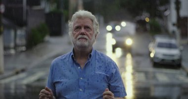 Yağmurda ayakta duran yaşlı adam, elleri açık ve duygusal ifade ile yukarı bakıyor keder, kafa karışıklığı, ya da ruhsal özlem şehir caddesinde