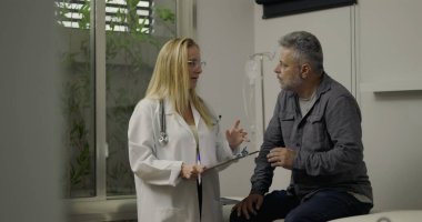 Klinikteki tıbbi randevu sırasında doktorla konuşan orta yaşlı erkek hasta, arka planda pano ve IV ekipmanıyla sağlık sorunlarını tartışıyor.