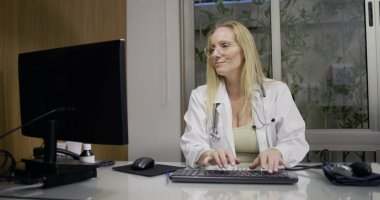 Tıp bürosunda masaüstü bilgisayarı üzerinde çalışan, hasta veri girişine odaklanan ya da klinik çalışma sırasında rapor yazan kadın doktor