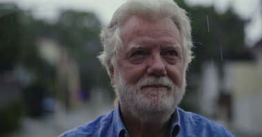 Yağmurda acı tatlı gülümseyen yaşlı bir adam, gözyaşı dolu gözler ve hüznü ve kabulü yansıtan şefkatli bir ifade.