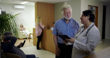Kadın doktoru dinlerken gülümseyen kıdemli adam klinik lobisindeki dijital tablet hakkında bilgi veriyor, resepsiyon masasının yanındaki günlük sağlık danışmanlığı