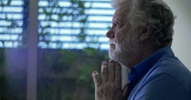 Yüzü nazik bir ifadeyle yukarı bakan yaşlı bir adam dua veya yansıma, elleri yavaşça birbirine yapışmış pencerenin yanında duruyor