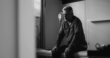 Klinikte başı dertteyken oturan orta yaşlı bir adam dramatik siyah-beyaz bir görüntü, tıbbi çevrede duygusal acı ya da zihinsel yük taşıyor.