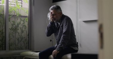 Orta yaşlı bir adam, boş bir muayenehanede gergin bir duruşla öne doğru eğiliyor. Zor bir muayeneden sonra, dışavurumunda görülen gerginlik ve yorgunluğu yansıtıyor.