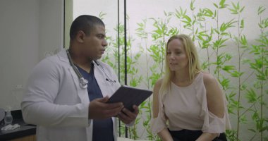 Klinik ziyareti sırasında orta yaşlı kadın hastaya tıbbi bilgileri açıklamak için tablet kullanan erkek doktor, sağlık hizmetlerinde güveni ve netliği artırıyor