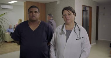 Hastane koridorunda yan yana duran sağlık personeli mesai sırasında kameraya tarafsız ifadelerle bakıyor.