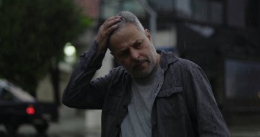 Orta yaşlı bir adam, yağmurun altında, elleri başının üstünde, gözleri açık, hayal kırıklığı ve zihinsel yorgunluk içinde.