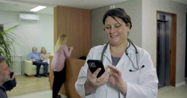 Hastanenin resepsiyonunda telefonla gülümseyen kadın doktor hastalar beklerken ve çalışanlar arka planda yürürken.