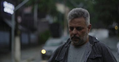 Orta yaşlı bir adam yağmurun altında duruyor. Islak giysiler ve kasvetli bir ifade, bulanık arabalar ve karamsar sokak atmosferiyle çevrili.