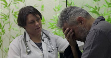 Sıkıntılı orta yaşlı erkek hasta yüzünü kapatırken, kadın doktor klinikte destek sunuyor, zor tıbbi haberlere duygusal ifadeyle tepki veriyor.