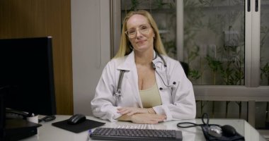 Klinikte çalışırken kameraya nazik bir ifadeyle bakan kadın doktor, masada tıbbi aletlerle oturuyor ve hesaplıyor.