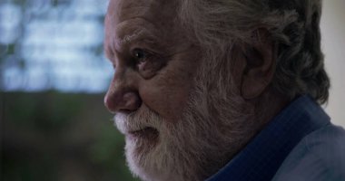 Yaşlı adam nazikçe gülümserken yumuşak arka plan mavisi ve kapalı mekan ortamında nazik ve düşünceli bir ifadeyle yan yana bakıyor.